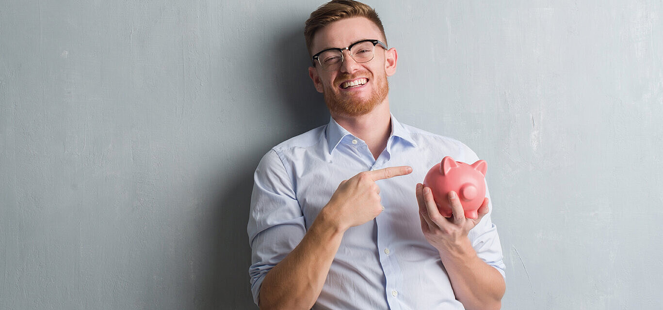 © Krakenimages - AdobeStock.com Smiling man pointing at piggy bank as a symbol of saving money