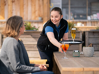 Beach Lounge Waitress serves Aperol Spritz and beer to a guest on the terrace of the Beach Lounge, with the beach in the background.