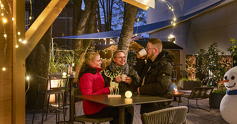 Maritim Christmas Village Three people enjoying hot drinks at the festive Christmas market of Maritim Hotel Darmstadt with fairy lights.
