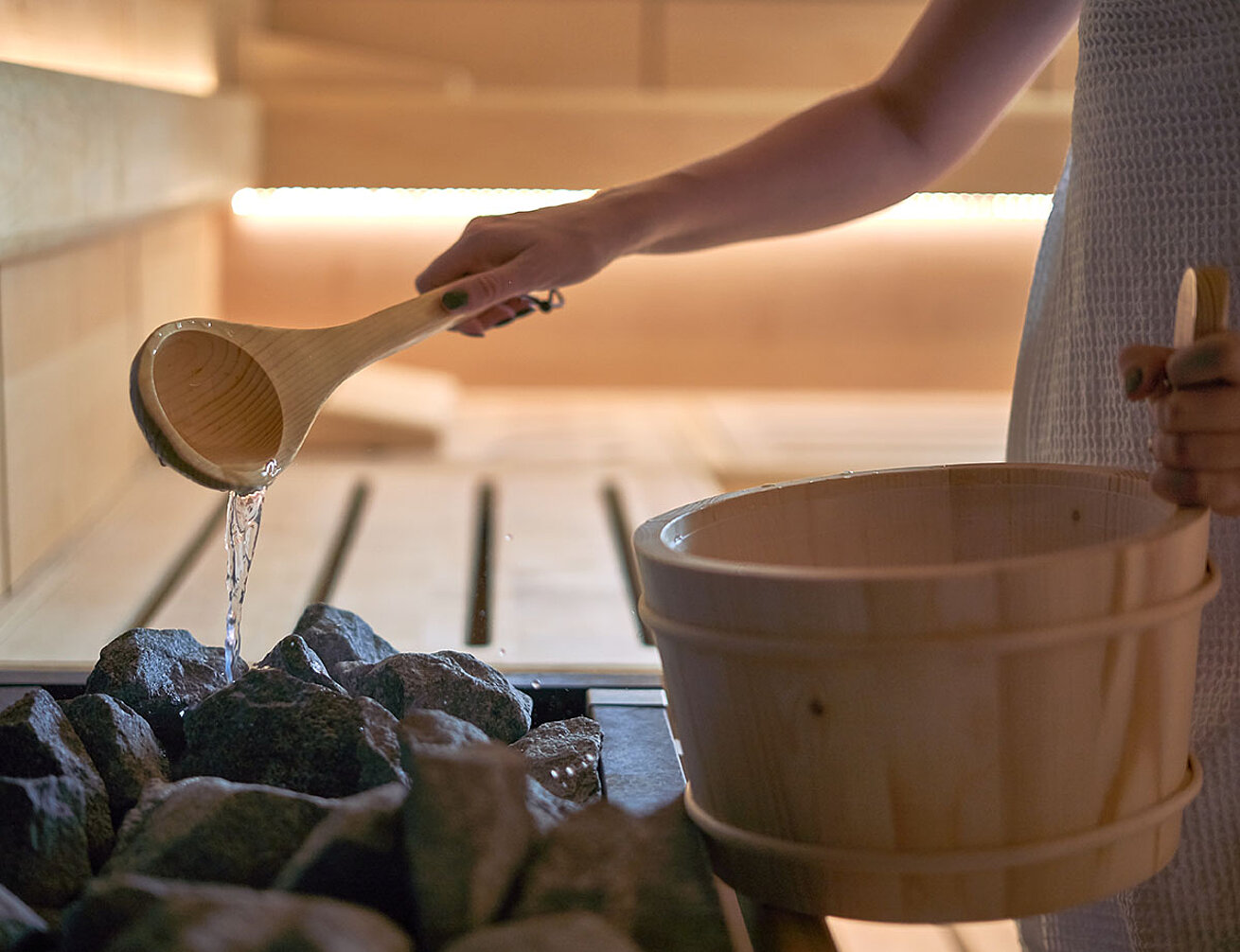 © a.dl - AdobeStock.com Person pouring water from wooden ladle onto hot sauna stones for infusion