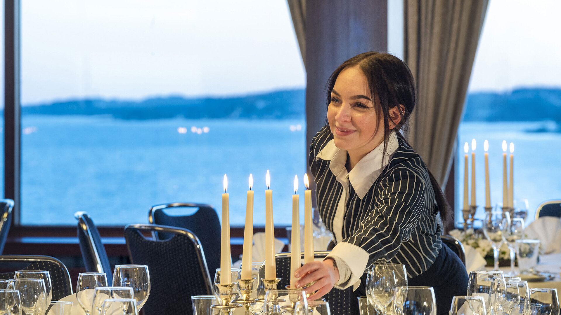 Maritim Conference Service Hall Maritim in the Maritim Hotel Kiel, a member of staff lights candles on a festively laid table, sea view in the background.