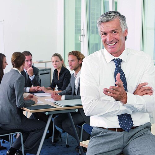 Mann im Businessmeeting Person in the foreground smiling while a business team works together at a meeting table in the background.