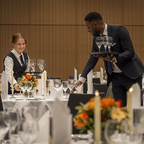 Service staff preparing tables in the Barbarossa Hall Service staff setting tables in the Barbarossa Hall of the Maritim Hotel Würzburg with floral decorations and glasses.
