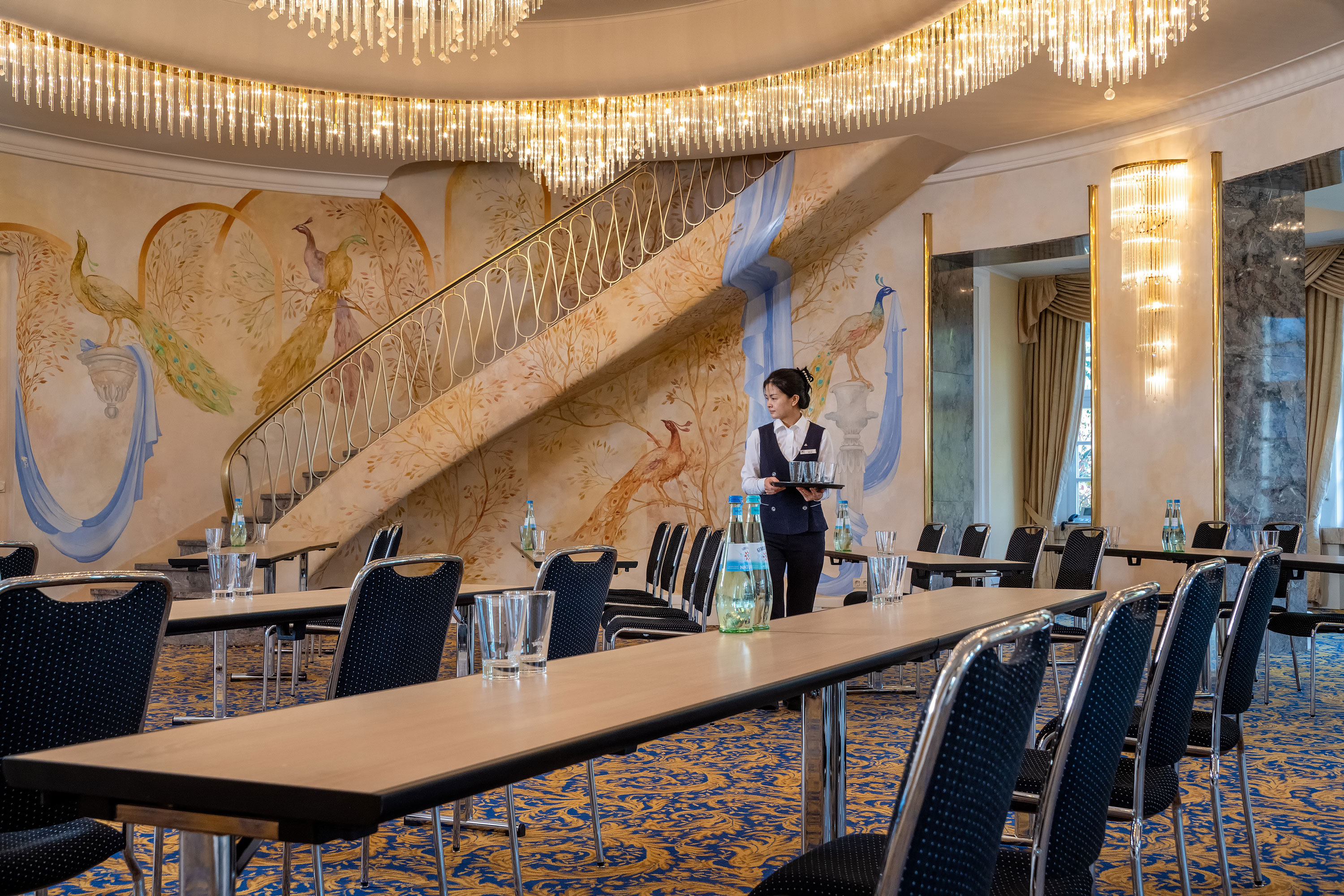 Fürst-von-Pückler-Saal Fürst-von-Pückler Hall with row seating, artistic wall design, large staircase and a staff member in the room.