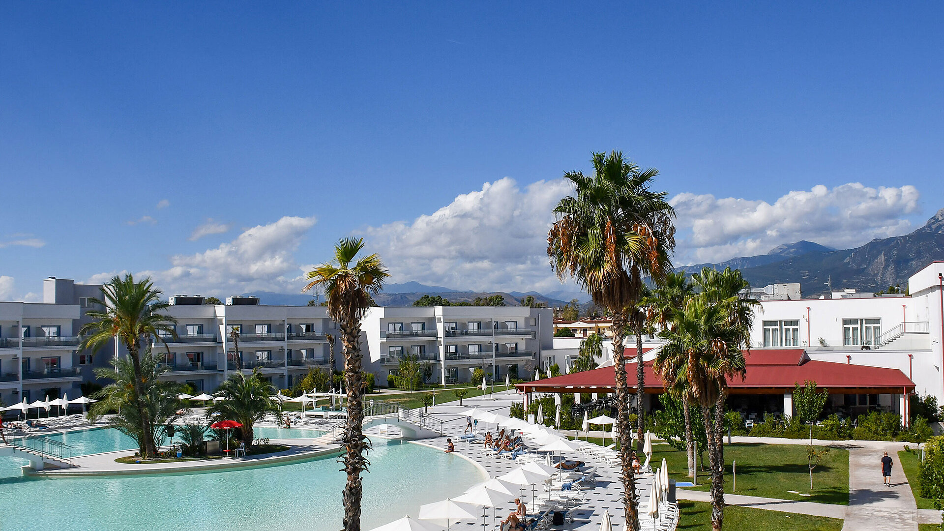 Exterior view Panorama of Maritim Resort Calabria with pool, palm trees, white buildings, and mountain backdrop under clear skies.