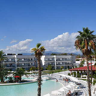 Panorama du Maritim Resort Calabria avec piscine, palmiers, bâtiments blancs et montagnes en arrière-plan sous un ciel clair.