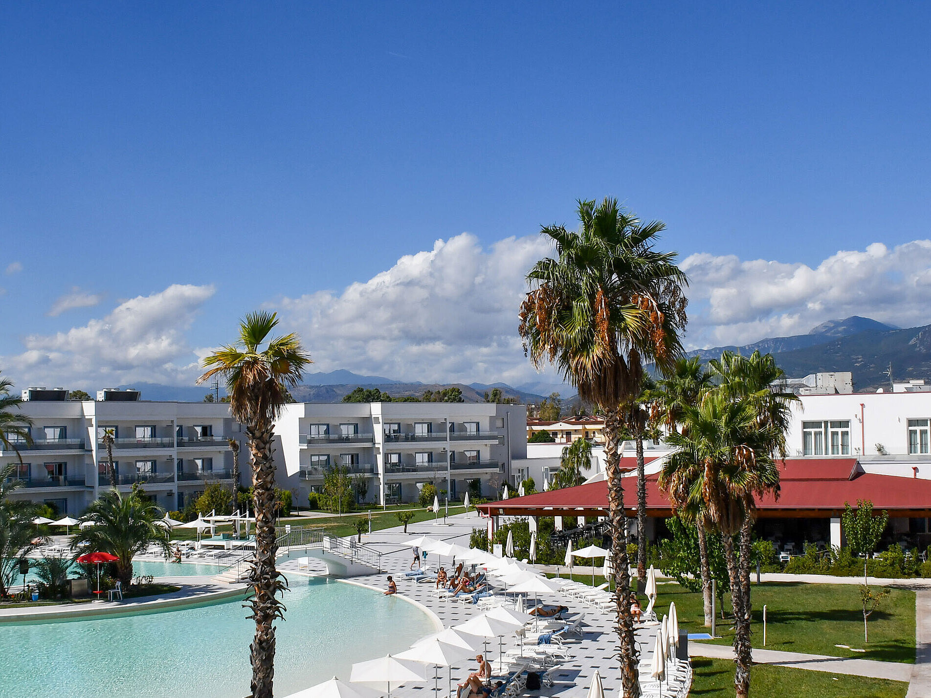 Exterior view Panorama of Maritim Resort Calabria with pool, palm trees, white buildings, and mountain backdrop under clear skies.