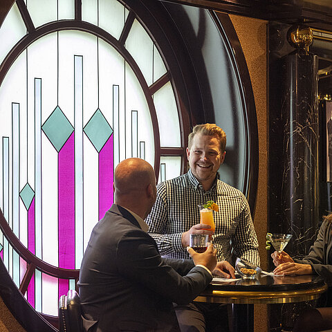 Drie gasten genieten van cocktails aan de pianobar in het Maritim Hotel Keulen, zittend voor een decoratief glas-in-loodraam.