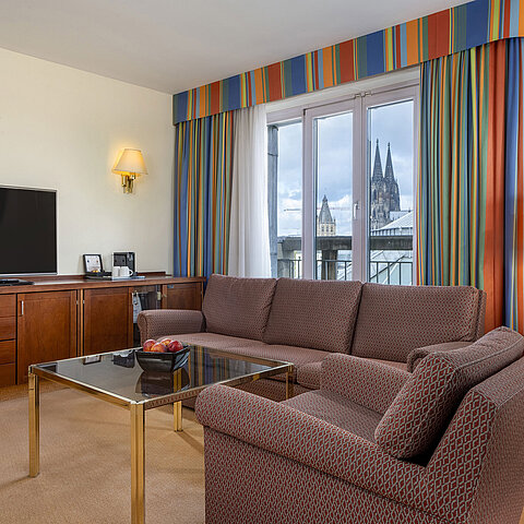 Soggiorno della Senior Suite al Maritim Hotel Köln con divano, TV e vista sul Duomo di Colonia.